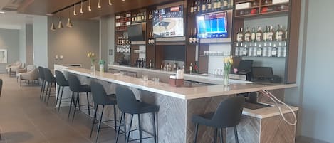 Zona de bar con una barra alargada, varios taburetes, una pantalla de televisión y estantes llenos de botellas.