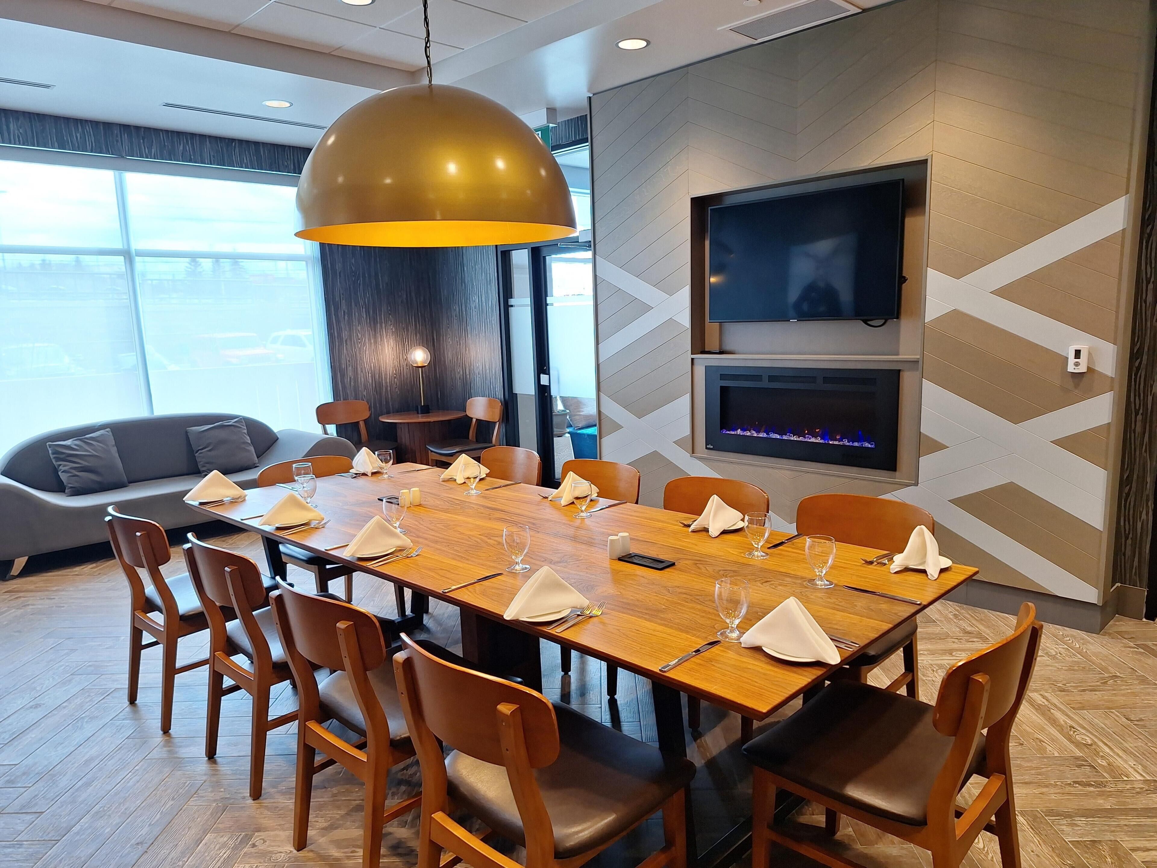 Un espace repas moderne avec une table en bois disposée pour accueillir six personnes, entourée de chaises en bois, d’un grand luminaire suspendu et d’un téléviseur à écran plat fixé au mur.