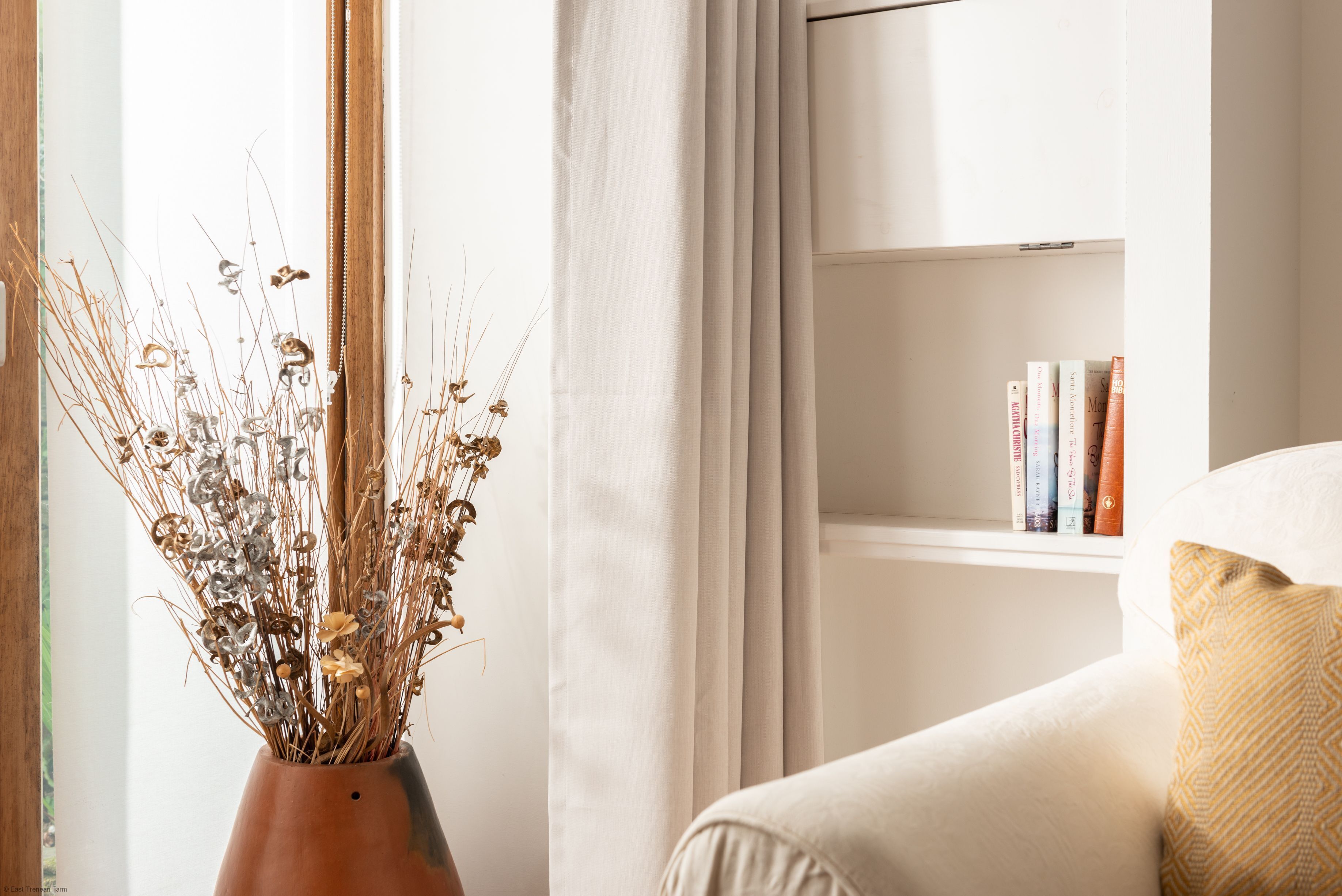 A room with a window, a bookshelf with books, a vase with dried plants, and a sofa with a cushion.