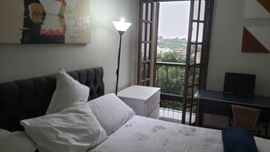 A bedroom with a bed, a desk, and a view of buildings through a window.
