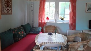 Ein gemütliches Zimmer mit einem roten Sofa, Korbstühlen, einem kleinen runden Tisch mit einer Vase und Kerze sowie einem Fenster mit Vorhängen.