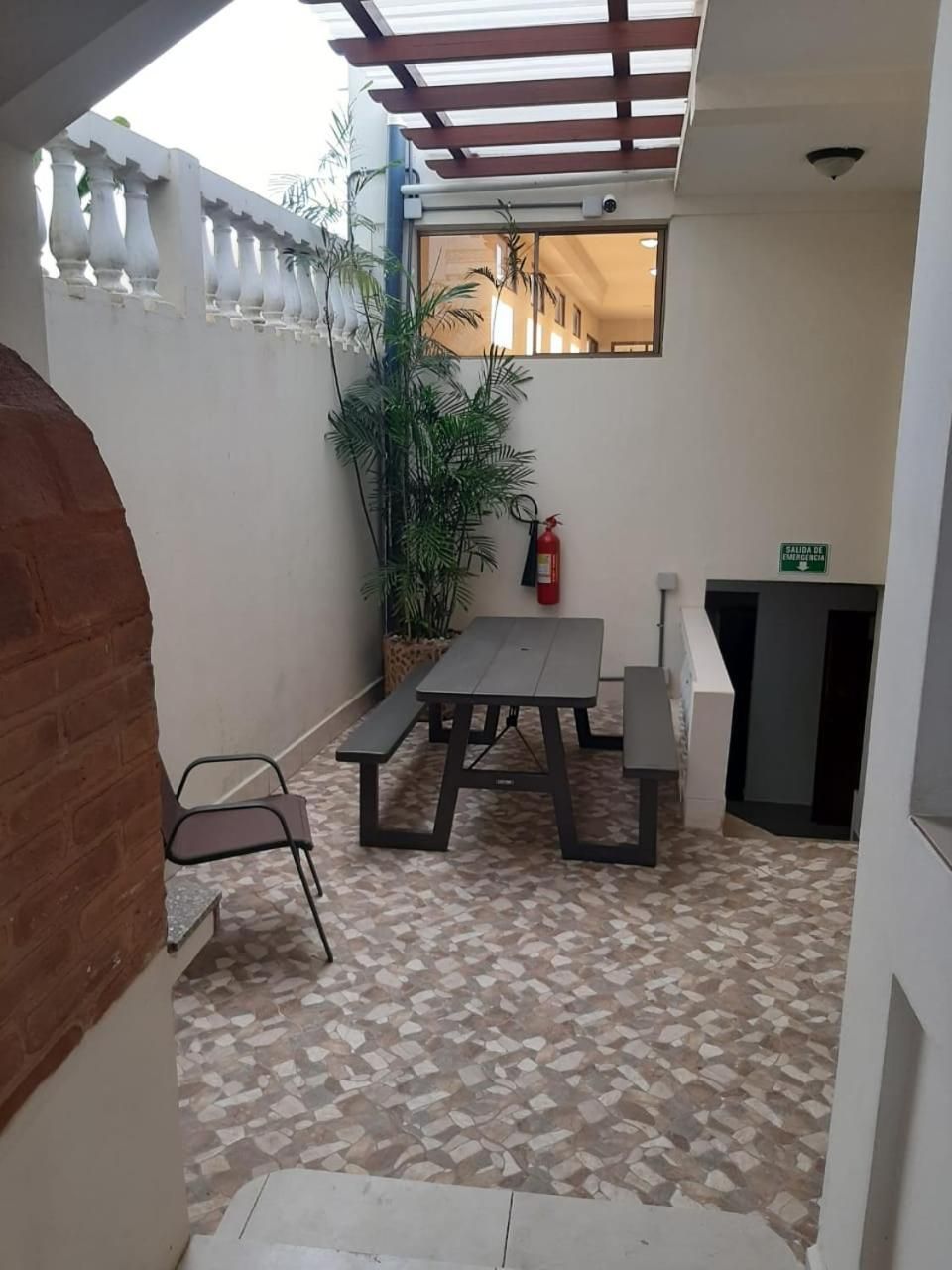 Área de pátio com mesa, bancos, extintor de incêndio, uma pequena planta e placa indicando saída de emergência.