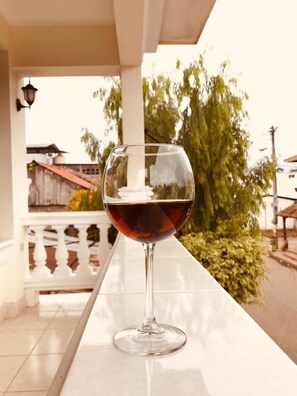 A glass of red wine on a white table outside a building with a white railing and a tree in the background.