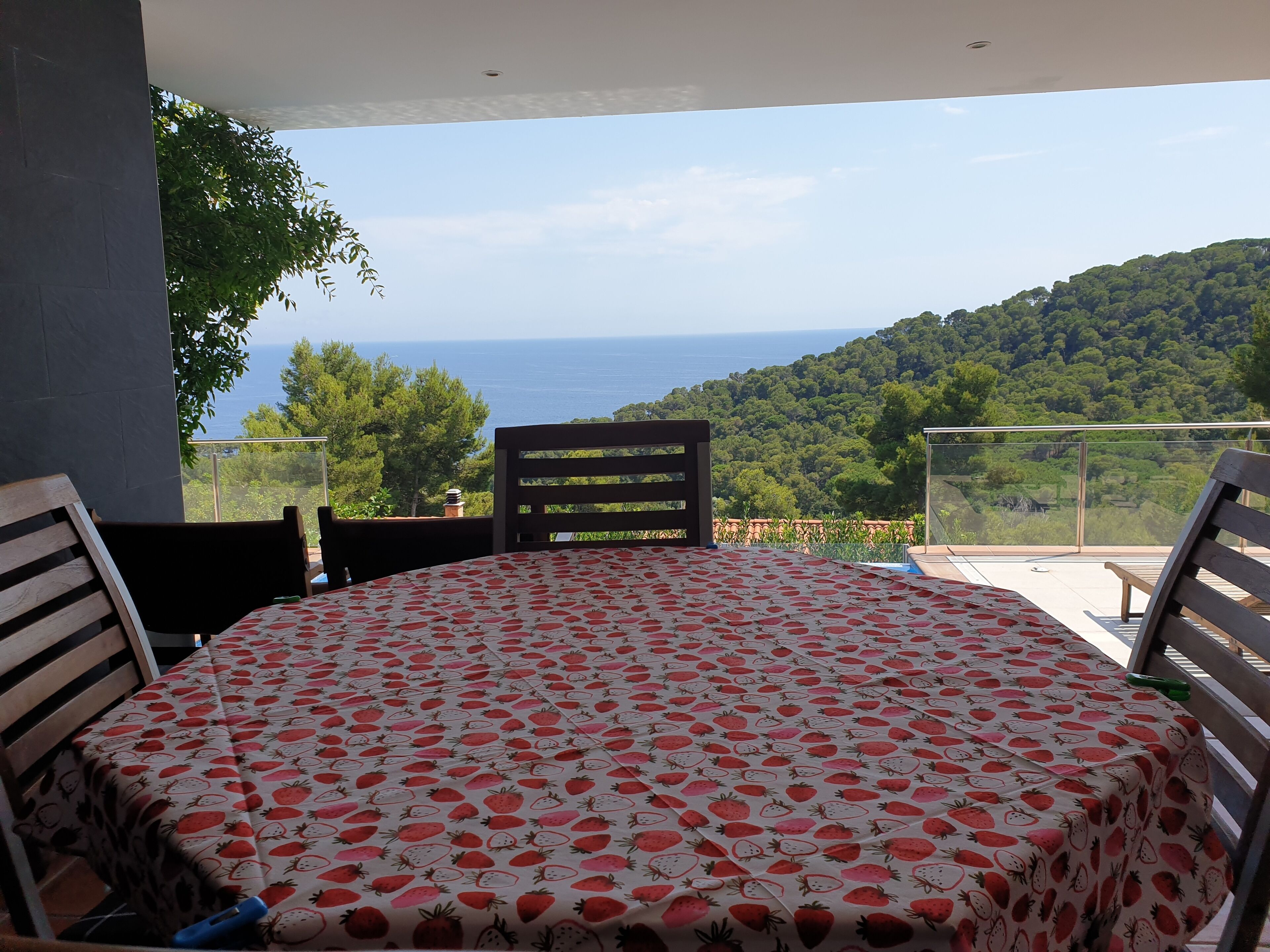 A round table with a strawberry pattern tablecloth, two wooden chairs, and a view of the sea and hills.