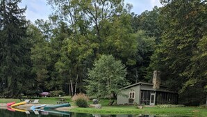 Une terrasse en bois avec une chaise et une tasse, donnant sur un étang paisible où flottent des bateaux, et avec une cabane visible au loin.