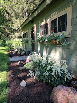 A house with a green exterior, a front porch with a door, and flower baskets on the windowsills. - Quaint Newly Remodeled Cabin with Stone Fireplace - Private Hot Tub - King Bed (Wooster)