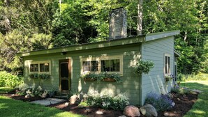 Une petite maison à un étage, à l’extérieur peint en vert, avec une cheminée en pierre et un jardin où poussent différentes plantes et où l’on trouve des rochers.