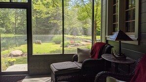 Une pièce dotée de grandes fenêtres donnant sur un jardin et un étang. On y trouve des meubles en osier et une petite table avec une lampe.