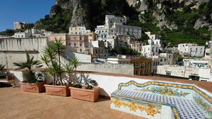Terraza en la azotea con macetas de terracota y una pared decorativa de azulejos, con vistas a un pueblo costero con edificios construidos en una ladera.
