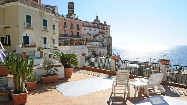 Una terraza en la azotea con plantas en macetas, mobiliario exterior y vistas al mar y edificios históricos.