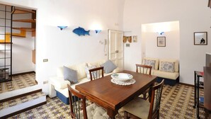 Una zona de comedor con una mesa y sillas de madera, un sofá con cojines estampados y suelo de baldosas.