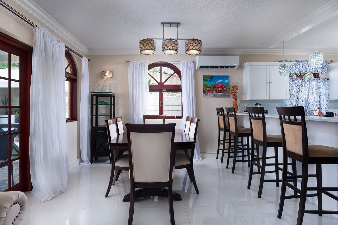A dining area with a long table, multiple chairs, and a kitchen area with a counter and hanging lights.