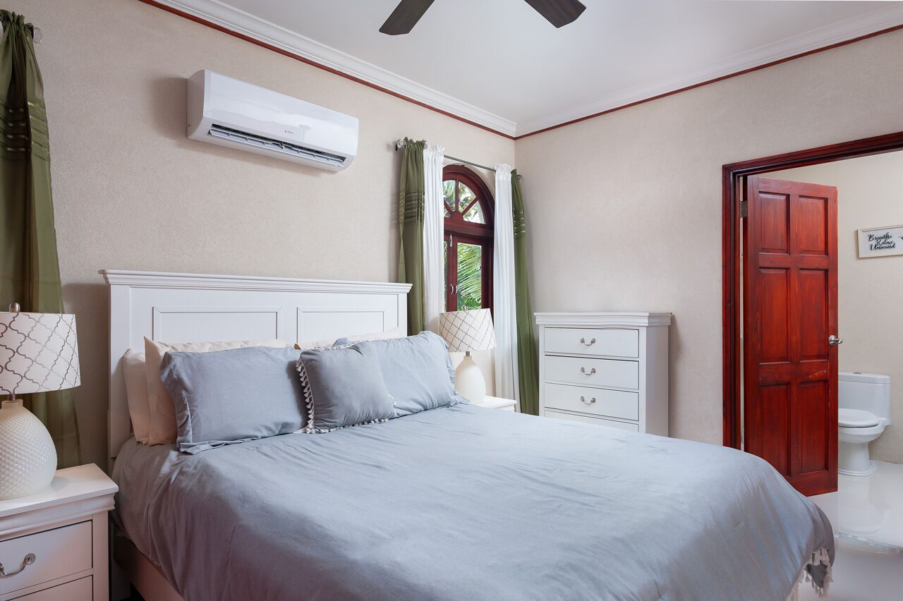 Une chambre à coucher comprenant un lit, une table de chevet, une commode, un appareil de climatisation, une fenêtre avec des rideaux et une porte.