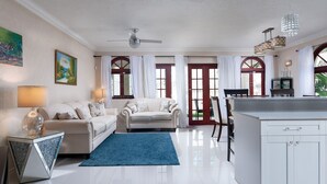 A spacious living room with a sofa, a coffee table, and a blue rug. There is a dining area with a table and chairs, and a kitchen area with cabinets and a countertop.
