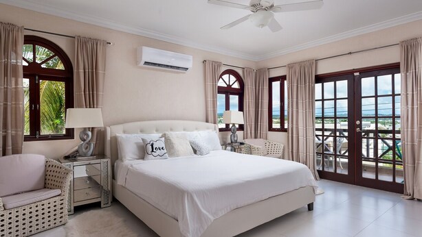 A spacious bedroom with a large bed, two bedside tables with lamps, a white sofa, and a view of the ocean through a sliding glass door.