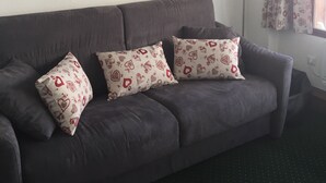 Een grijze bank met gekleurde kussens, een ronde spiegel en een raam met gordijnen met een patroon.