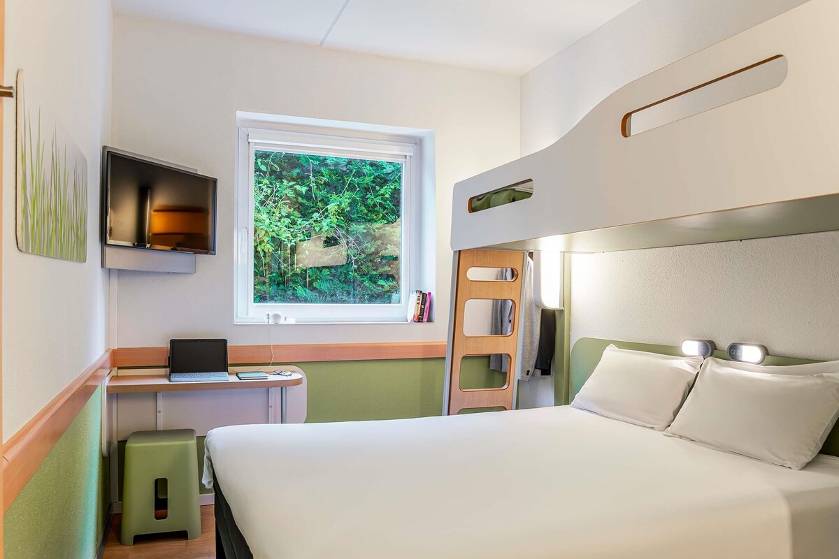 A hotel room with a bed, a desk with a laptop, a window with a view of greenery, and a small stool.