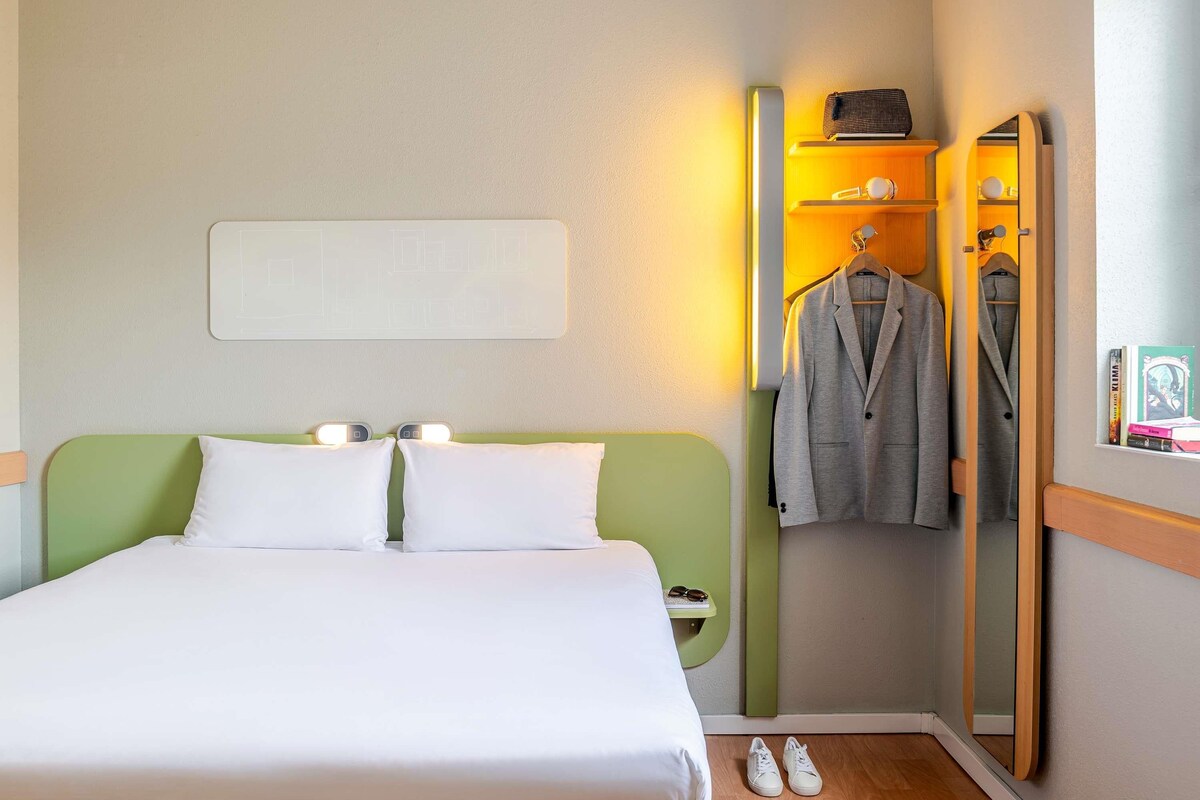 A hotel room with a bed, bedside lamps, a mirror, a coat rack, and a small shelf with items.