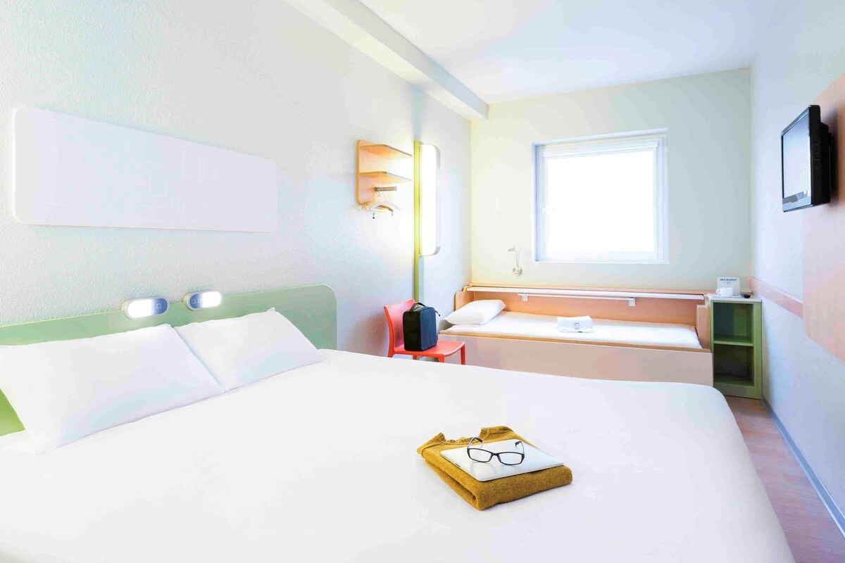 Una habitación de hotel con cama, ventana, mesita de noche y televisión.