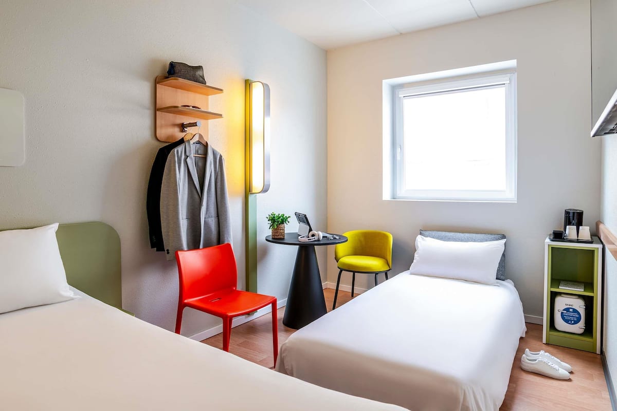 Habitación de hotel con dos camas, una silla roja, una silla amarilla, una mesita con una planta y una ventana.