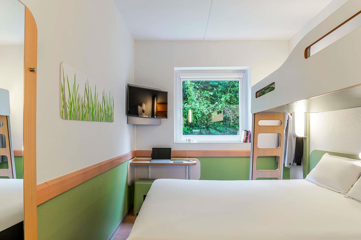 Habitación de hotel con cama, escritorio con una computadora portátil, televisor y ventana con vista a follaje.
