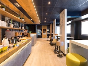 A modern cafe interior with a wooden counter, seating area, and a vending machine. - Ibis Budget Bilbao Barakaldo (Barakaldo)
