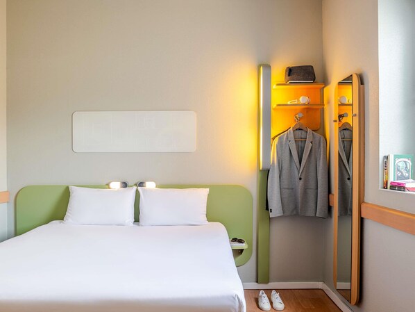 Habitación de hotel con cama, lámparas de noche, espejo, perchero y un estante con objetos.
