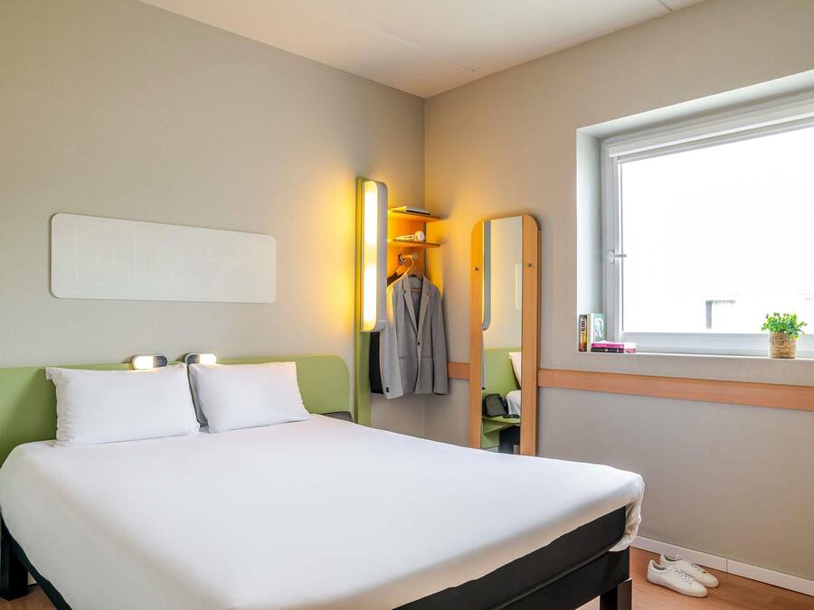 Habitación de hotel con cama, mesita de noche, espejo, ventana y lámpara de pared.