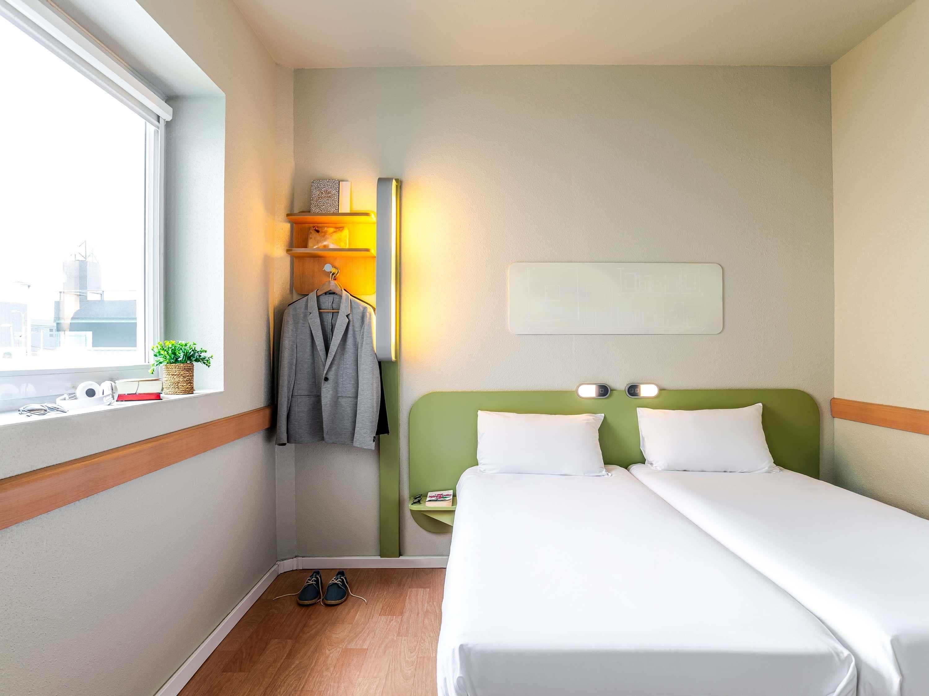 Habitació d'hotel amb un llit, una taula de nit, una làmpada, una finestra amb vistes i un penjador amb una jaqueta.