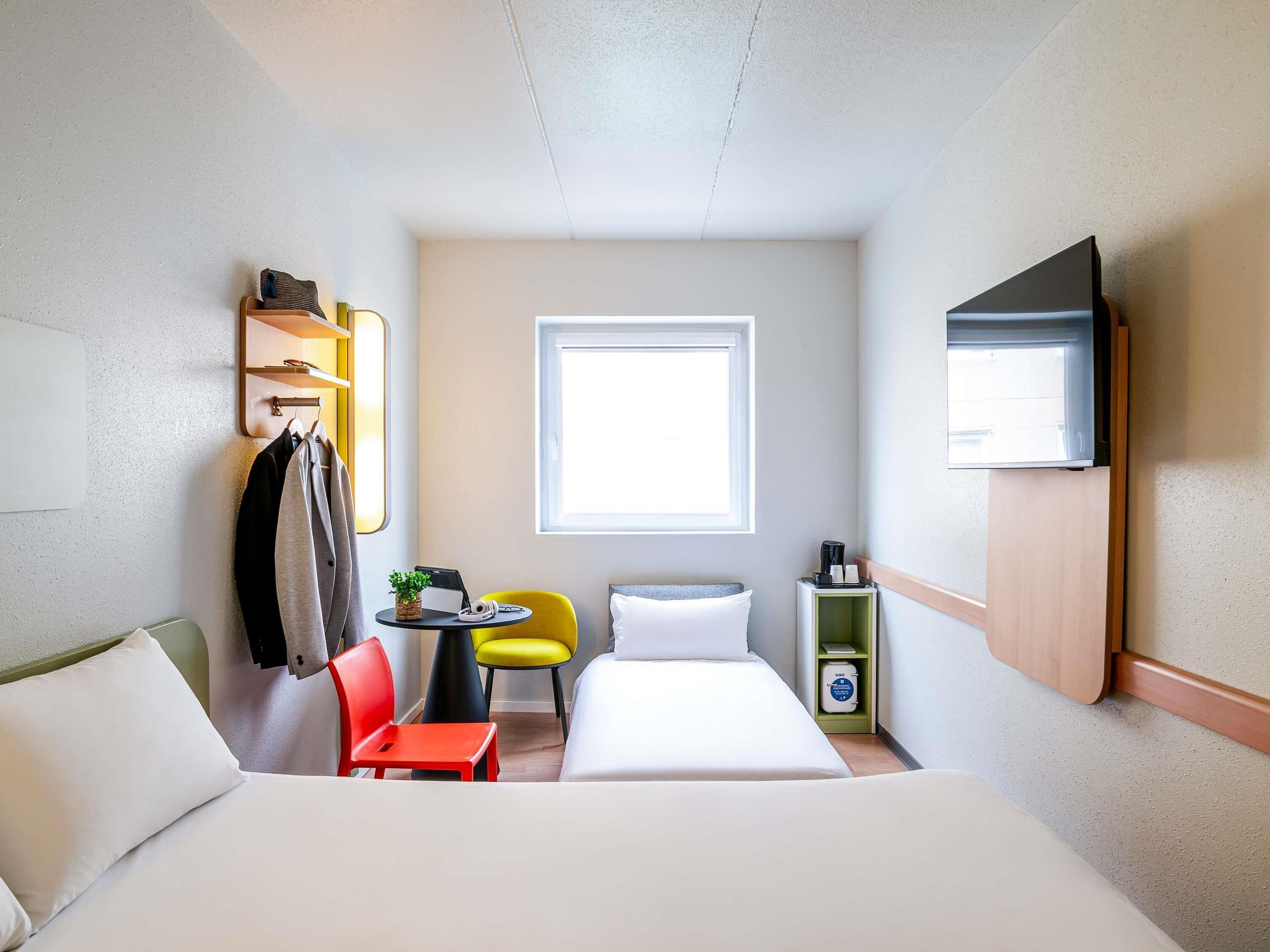 A compact hotel room with a bed, a desk with a chair, a small table with a plant, a window, and a TV mounted on the wall.