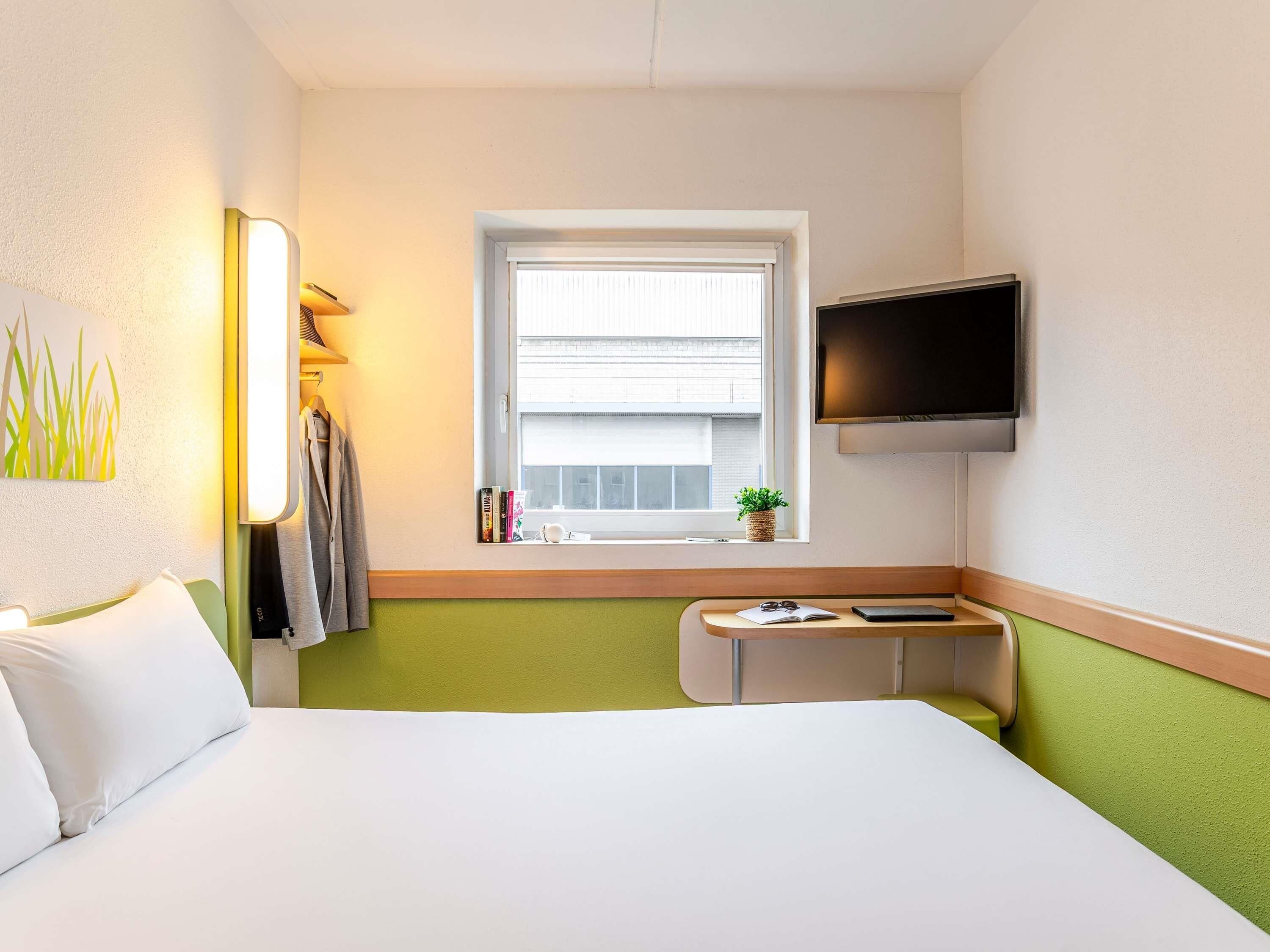 A hotel room with a bed, a desk, a TV, a window with a view, and a plant on the windowsill.
