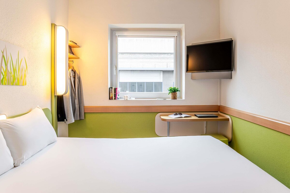A hotel room with a bed, a desk, a TV, a window with a view, and a plant on the windowsill.