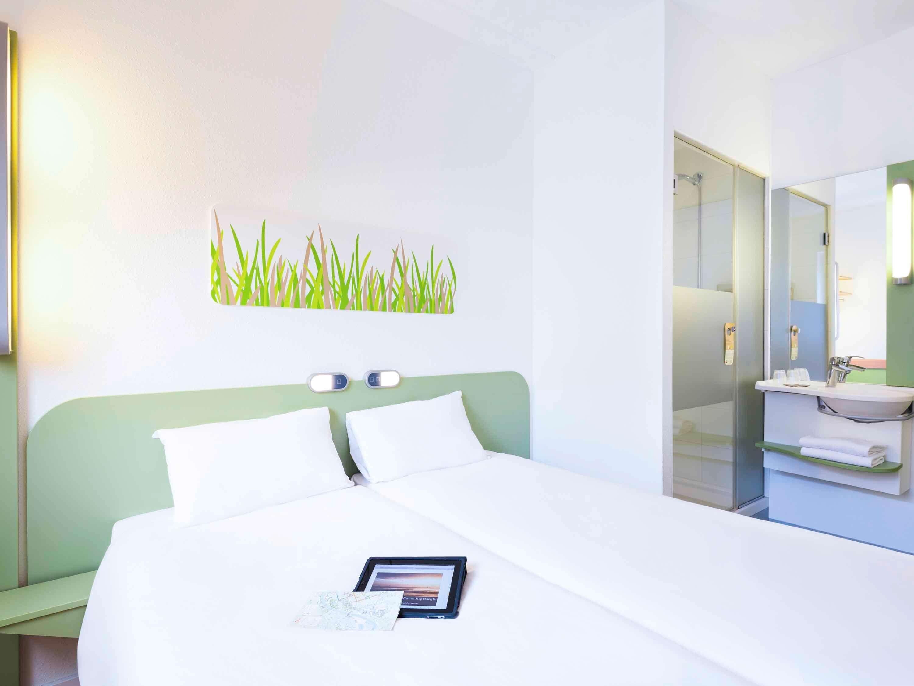 A hotel room with a bed, a bedside table, a nightstand, a wall-mounted light, a mirror, and a bathroom with a sink and towel rack.
