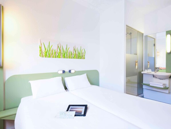 A hotel room with a bed, a bedside table, a nightstand, a wall-mounted light, a mirror, and a bathroom with a sink and towel rack. - Ibis Budget Bilbao Barakaldo (Barakaldo)