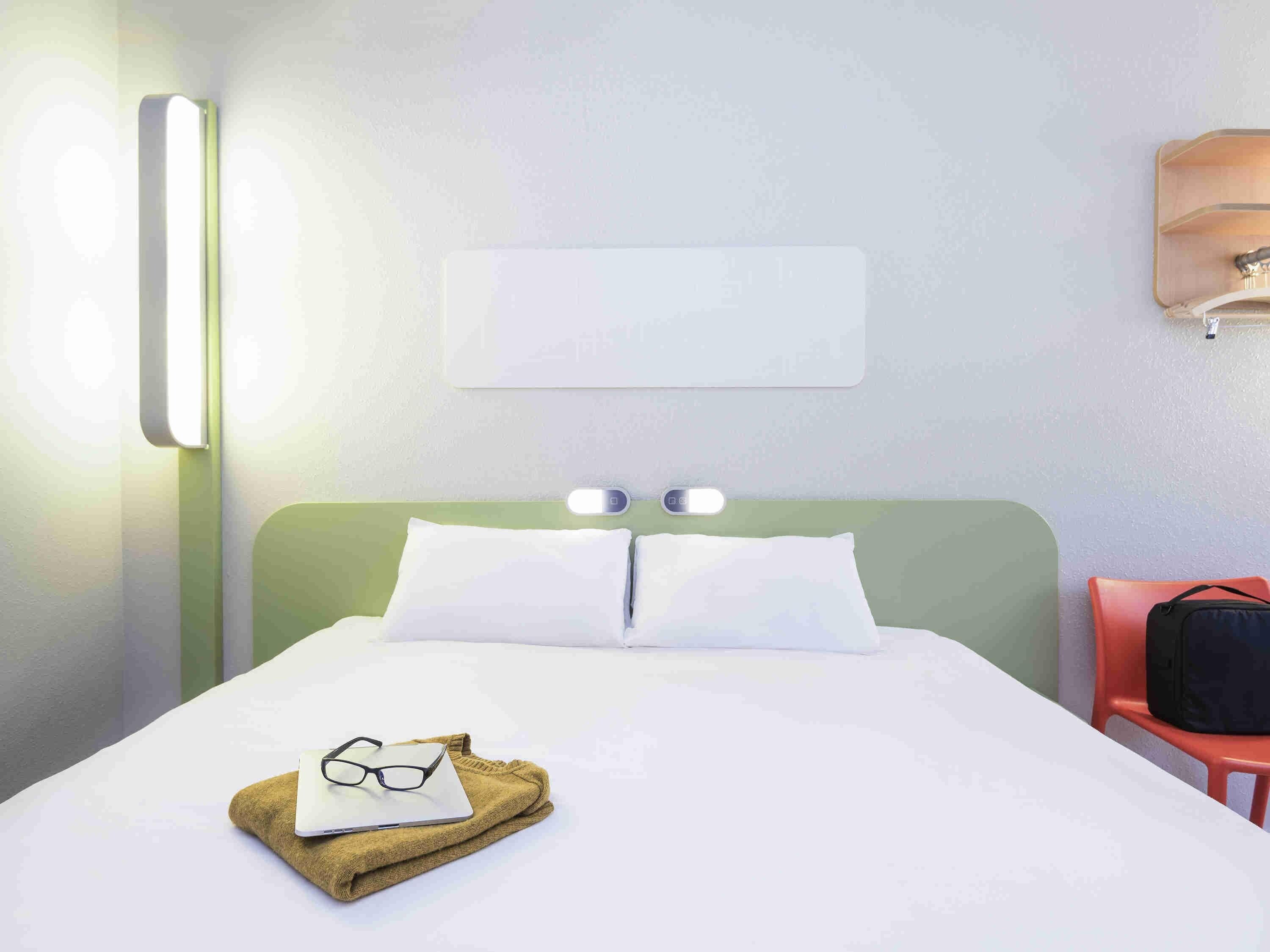 A hotel room with a bed, a nightstand, a wall-mounted lamp, and a chair.