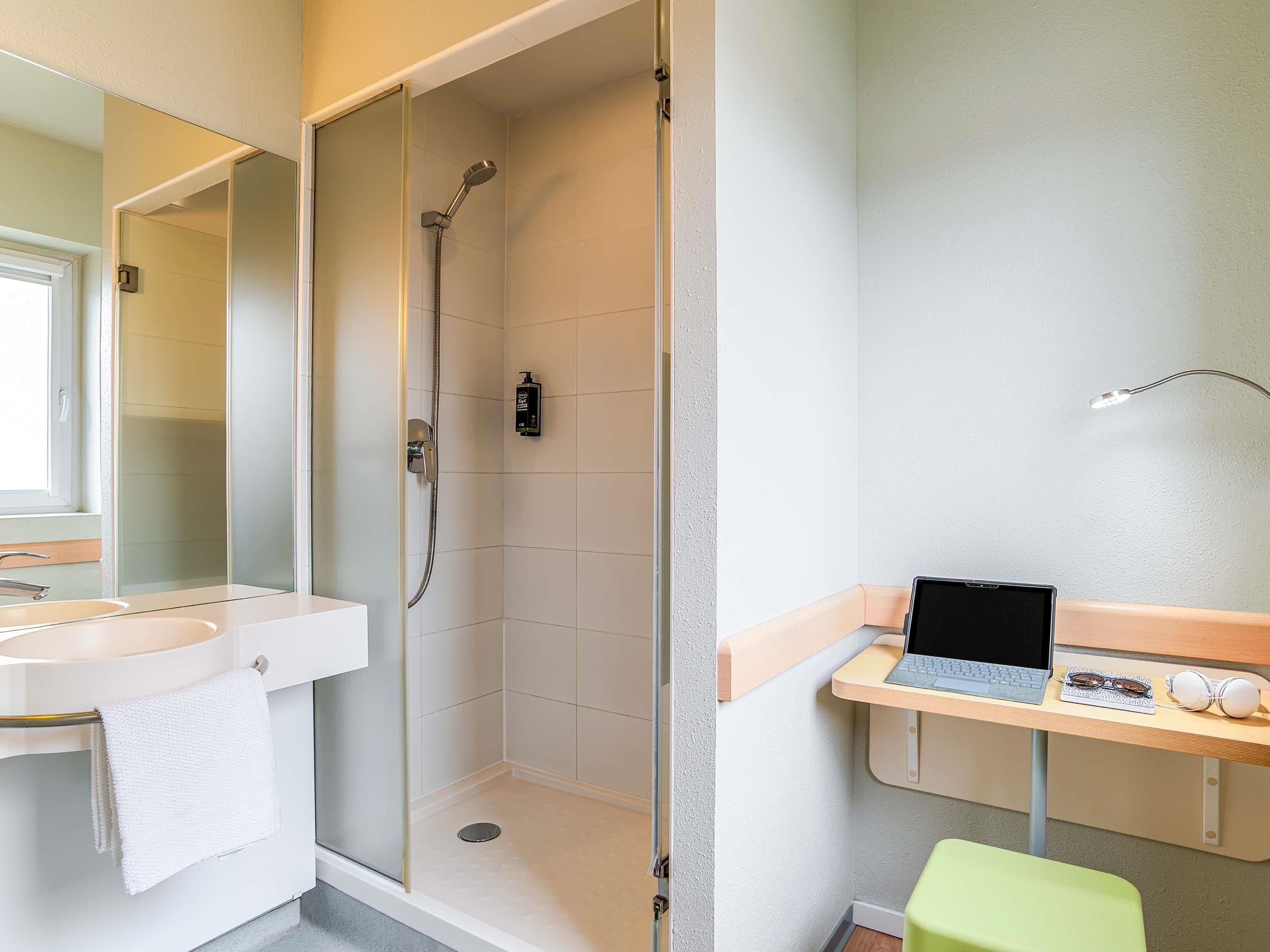 Ein Badezimmer mit Dusche, Waschbecken, Spiegel und einem Schreibtisch mit Laptop, Brille und Kopfhörern.