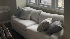 A room with a white sofa, two pillows, and a window with a view of a street and buildings.