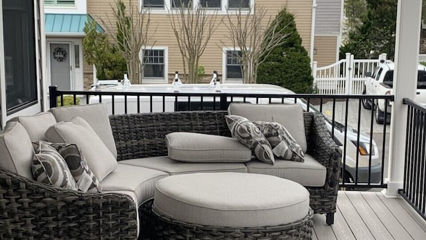 A covered outdoor seating area with a sectional sofa and ottoman, a wicker chair, and a wooden deck.