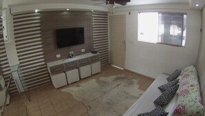 Ein modernes Wohnzimmer mit Fernseher, Deckenventilator, einer Couch mit gemusterten Kissen und einem großen Fenster mit Jalousien.
