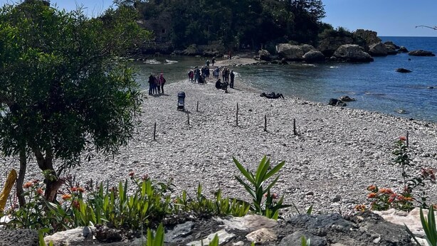 Une plage rocheuse où se trouve un petit groupe de personnes, un ciel dégagé et, au loin, une colline boisée.