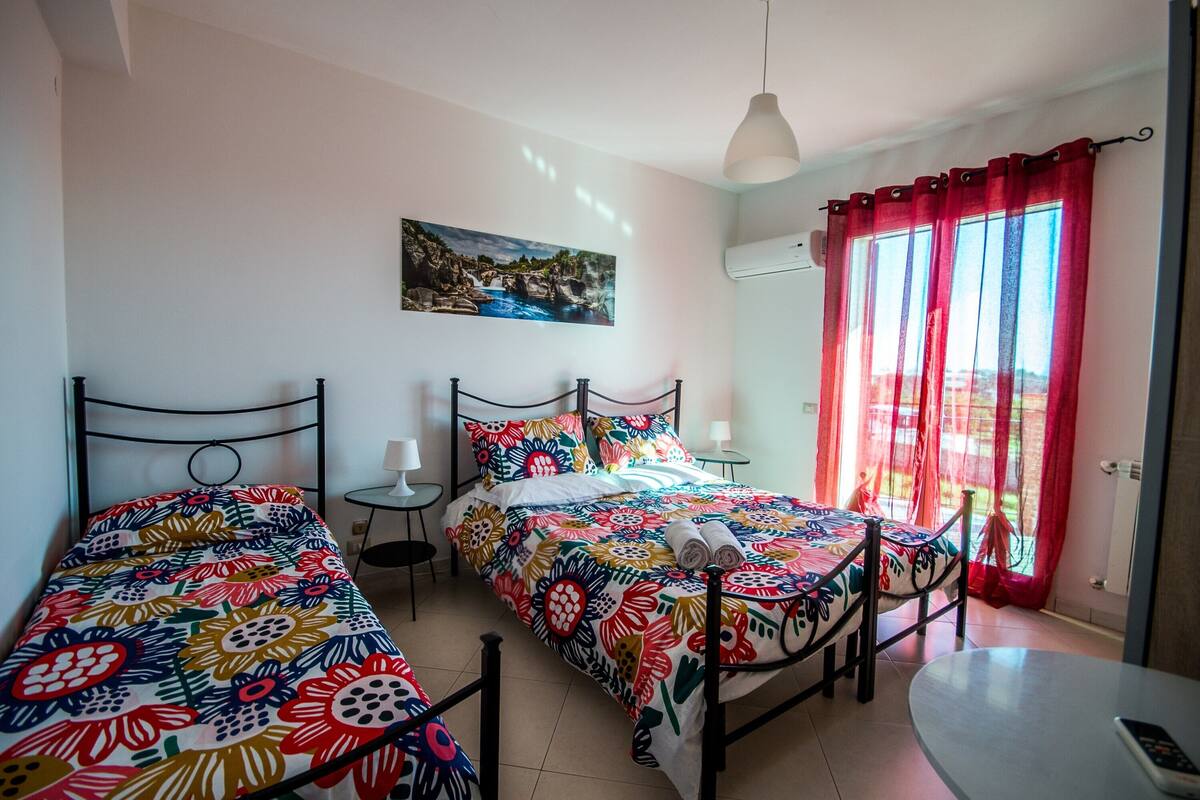 Une chambre avec deux lits, une table ronde, un tableau au mur et une fenêtre avec des rideaux rouges.