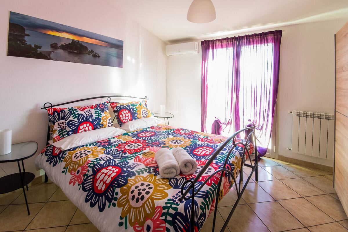 Une chambre à coucher avec une couvre-lit à motifs floraux aux couleurs vives, une table de chevet, une chaise et une vue sur la mer par une fenêtre ornée de rideaux violets.