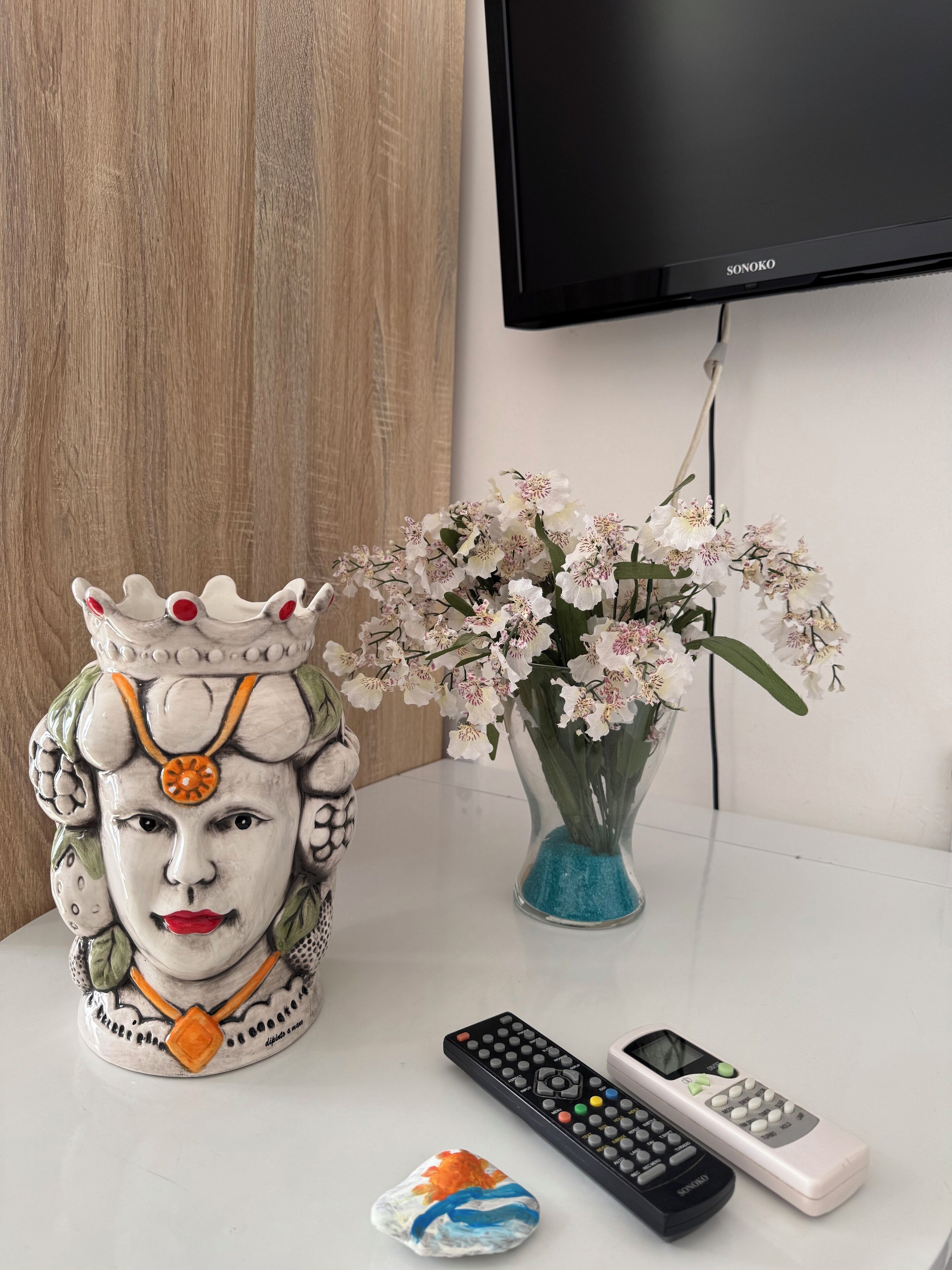 A ceramic figure with a crown, a vase with white flowers, and two remote controls on a white surface.