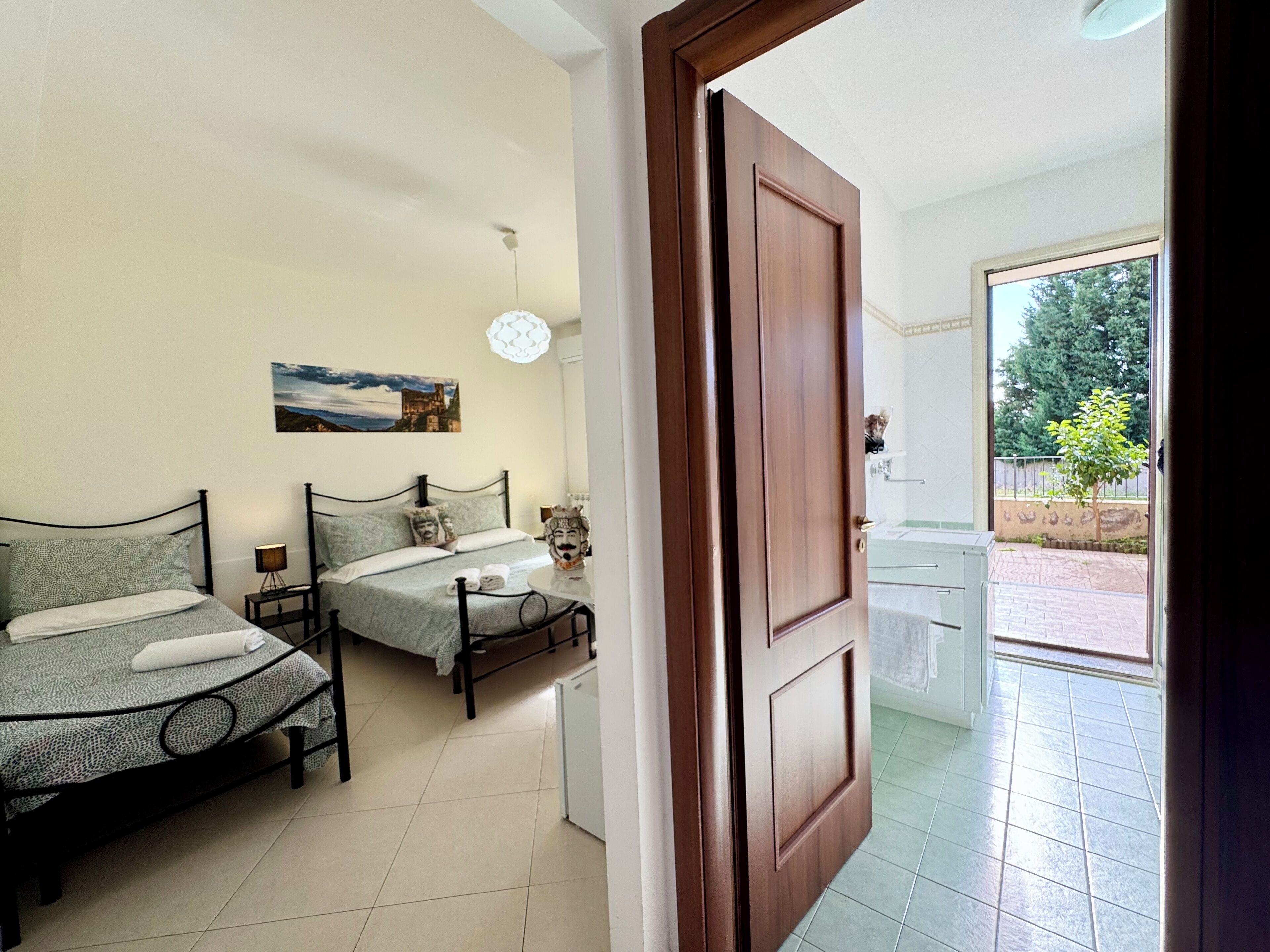 A bedroom with two beds, a painting on the wall, and a view of the outside through an open door.