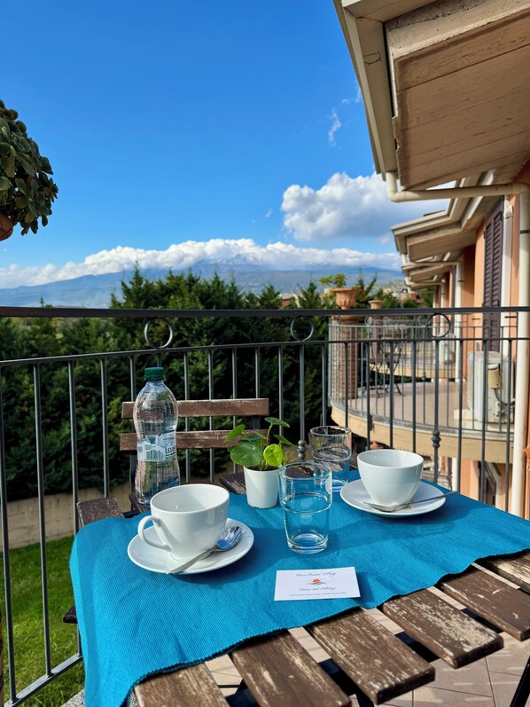 Mesa en balcón con tazas blancas, una botella de agua y una pequeña planta, con vistas a un paisaje montañoso.