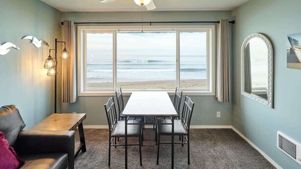 A room with a dining table and chairs, a ceiling fan, a large window with a view of the ocean, and a decorative mirror.