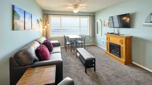 A living room with a sofa, a coffee table, a fireplace, a TV, and a large window with a view of the ocean.