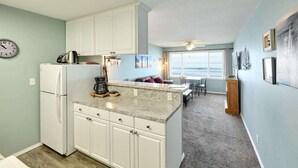 A compact kitchen with white cabinets, a granite countertop, and a refrigerator. There is a clock on the wall, and a dining area with a view of the ocean.