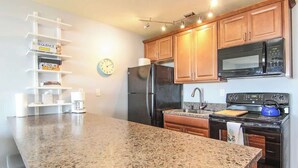 Una cocina con encimeras de granito, armarios de madera, refrigerador de acero inoxidable, microondas, horno y hervidor.