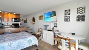 Una habitación de hotel compacta con kitchenette, una cama con edredón con motivos marinos azules, una zona de comedor con mesa y sillas blancas y un televisor montado en la pared.